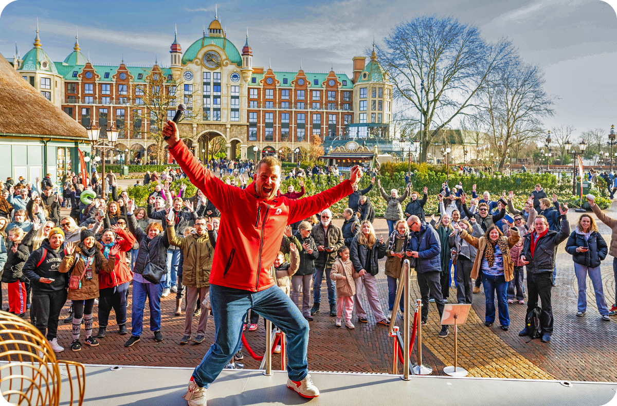 Een presentator staat enthousiast voor een grote, vrolijke groep mensen op een plein in attractiepark de Efteling te zien.