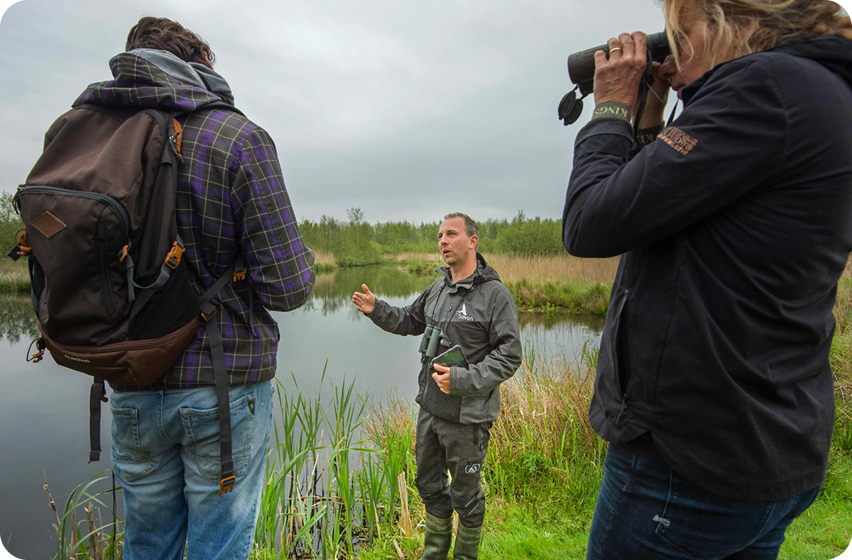 Een medewerker van Sovon geeft uitleg aan twee bezoekers bij een meer omringd door gras, riet en andere planten.