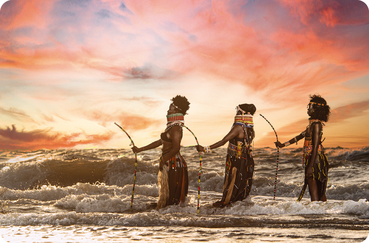 Drie vrouwen in traditionele kleding staan met handbogen in de zee bij een kleurrijke zonsondergang.