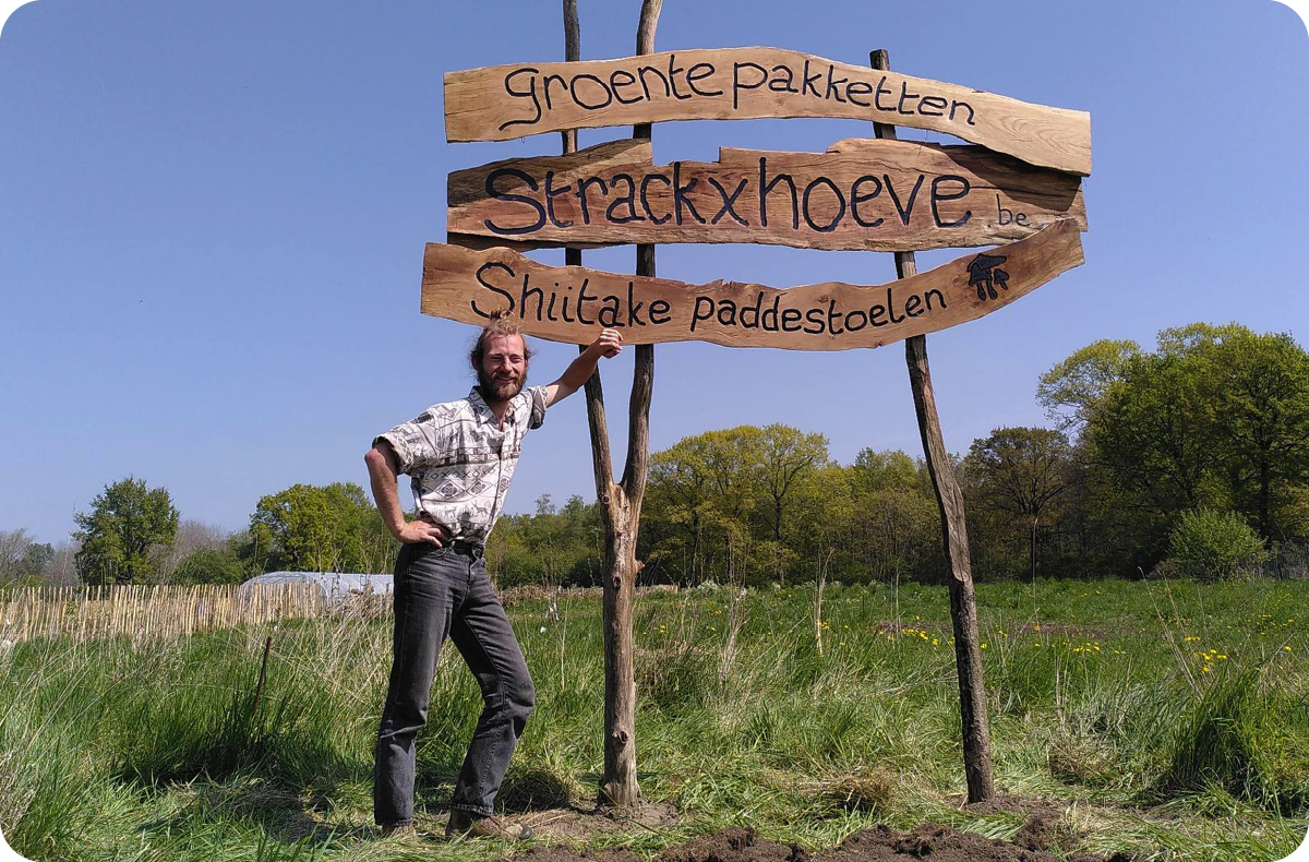 Een man staat in een grasveld naast een houten bord met de tekst: Groentepakketten, Strackxhoeve.be en Shiitake paddenstoelen.