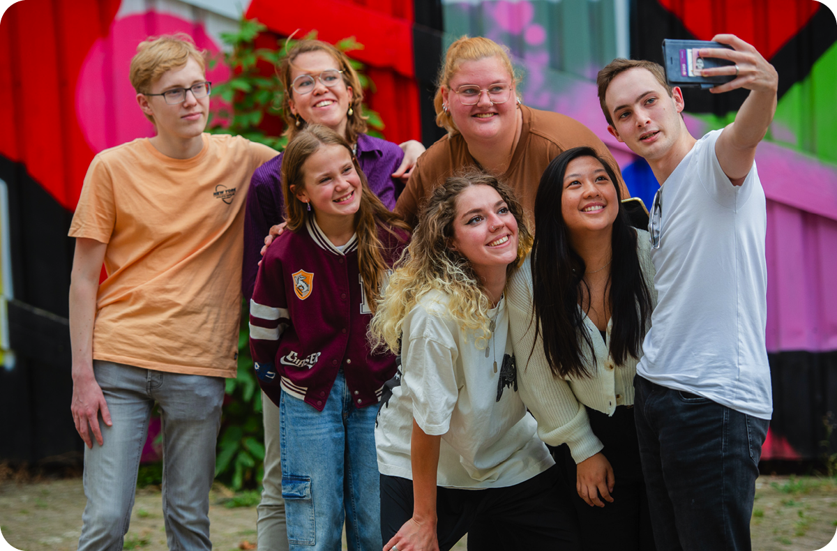 Een groep van zeven jongeren die samen poseren voor een selfie voor een kleurrijke muur met graffiti.