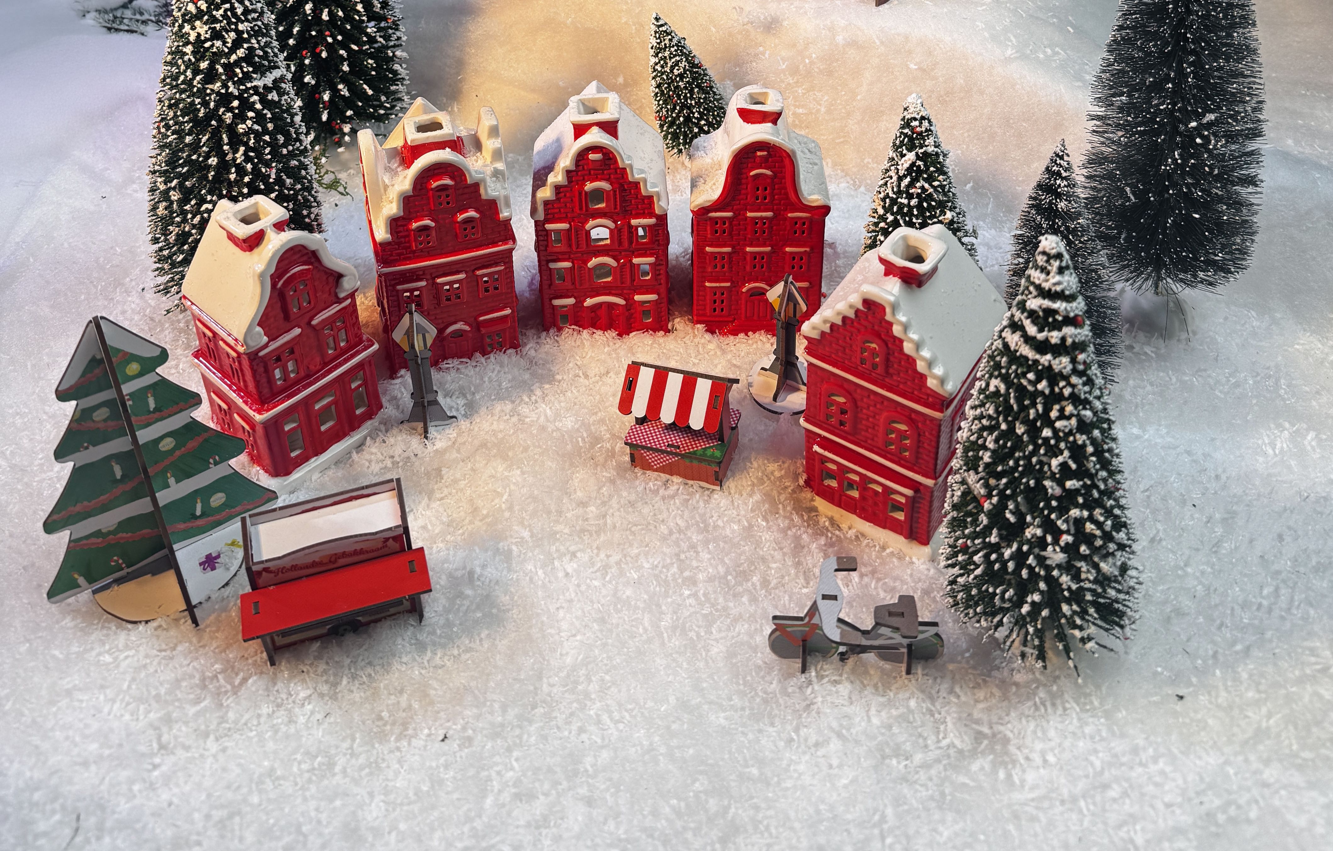 Miniatuur winters dorp met vijf rode huizen, groene bomen, een bankje, een fietsje en een marktkraam in de sneeuw.